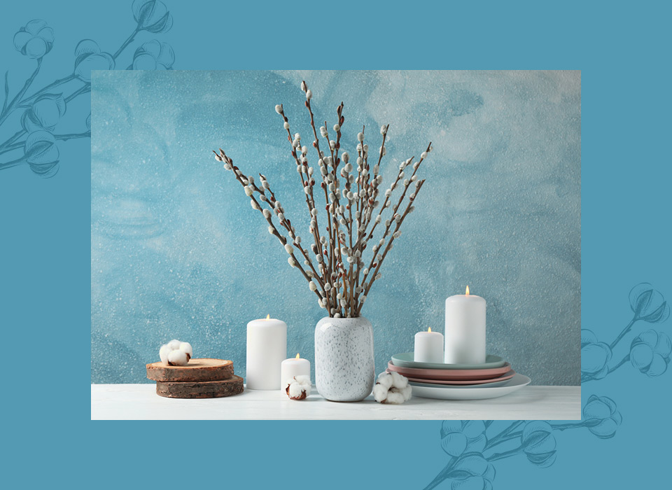 Arranjo decorativo com velas brancas acesas, vaso com galhos ornamentais e elementos naturais sobre fundo azul suave, criando uma atmosfera elegante e acolhedora.
