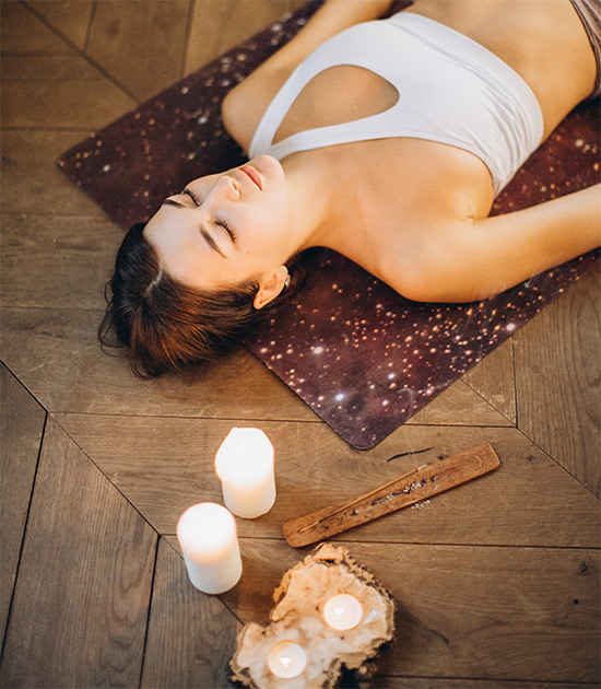 Pessoa relaxando sobre um tapete de yoga enquanto velas acesas e incenso criam uma atmosfera tranquila e acolhedora em ambiente interno.