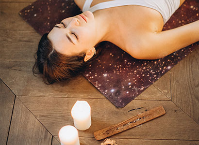 Pessoa relaxando sobre um tapete enquanto velas acesas e incenso criam uma atmosfera tranquila e acolhedora para meditação e bem-estar.