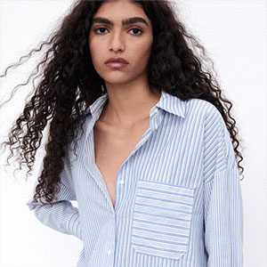 Pessoa com cabelos longos e cacheados usando camisa listrada azul e branca, em retrato de estilo editorial sobre fundo claro.