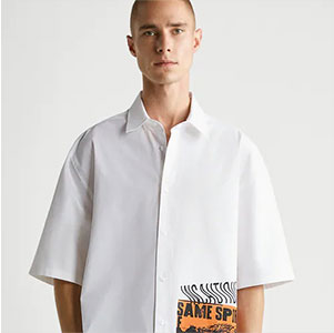Pessoa usando camisa branca de modelagem ampla com detalhes gráficos discretos, posando diante de um fundo neutro em estilo editorial contemporâneo.