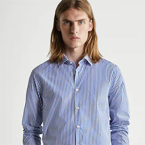 Pessoa com cabelos longos usando camisa social listrada em azul e branco, posando em frente a um fundo neutro em estilo editorial de moda.
