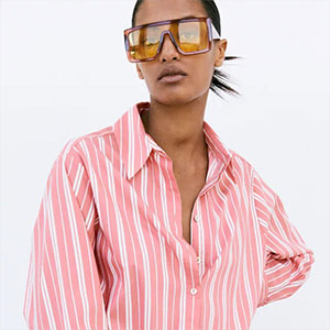 Pessoa usando camisa listrada em tons de rosa e branco e óculos de sol com lentes coloridas, em retrato de estilo editorial sobre fundo claro.