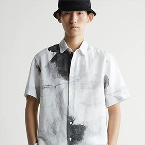Pessoa usando chapéu bucket preto e camisa branca com estampas artísticas em tons escuros, posando diante de um fundo neutro em estilo editorial.