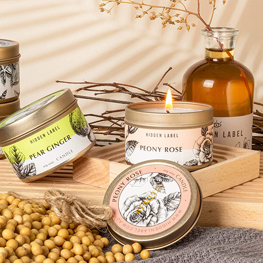 Conjunto de velas aromáticas em latas metálicas decoradas, dispostas sobre bandeja de madeira com elementos naturais, galhos secos, sementes e frasco de vidro decorativo.