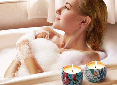 Mulher relaxando em uma banheira com espuma enquanto duas velas aromáticas decorativas acesas estão posicionadas na borda.