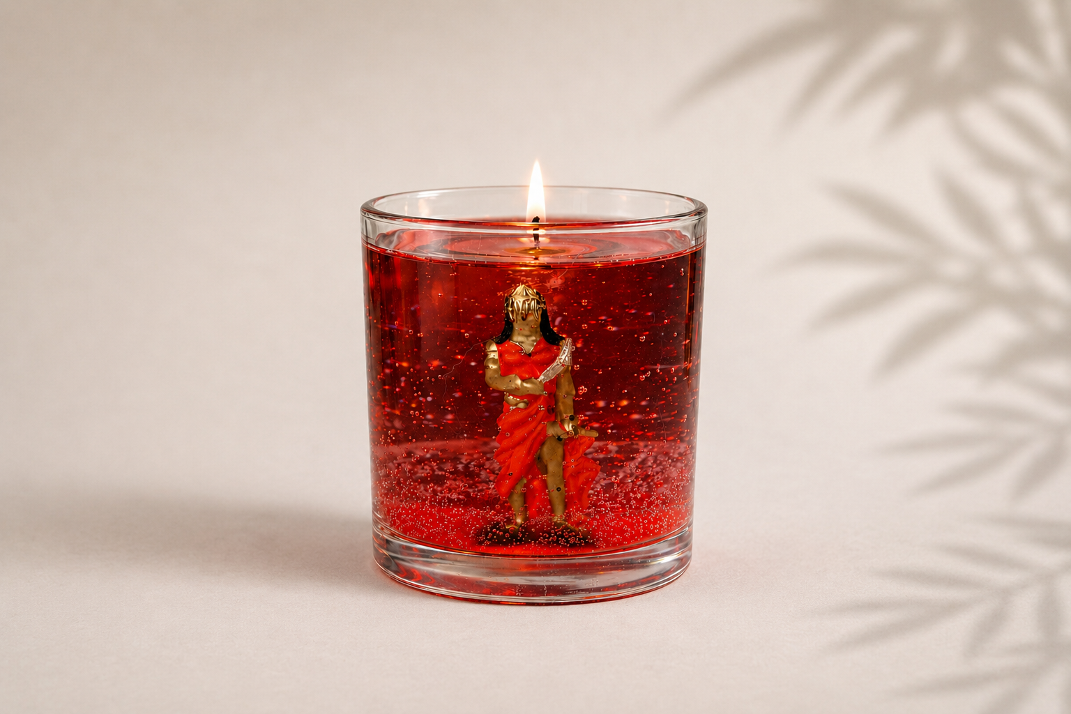 Vela em Gel Iansã artesanal em copo de vidro redondo com parafina em gel vermelha translúcida, contendo uma representação de Iansã em seu interior. A vela está acesa e posicionada sobre fundo claro com iluminação suave e sombras delicadas de folhagens.