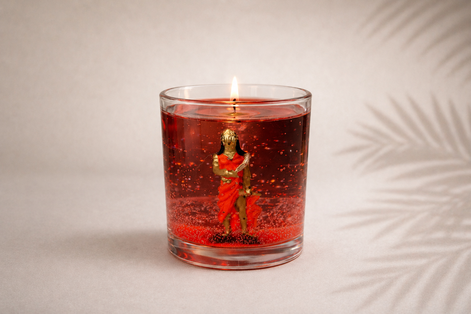 Vela em Gel Iansã artesanal em copo de vidro redondo com parafina em gel vermelha translúcida, contendo uma representação de Iansã em seu interior. A vela está acesa e posicionada sobre fundo claro com iluminação suave e sombras delicadas de folhagens.