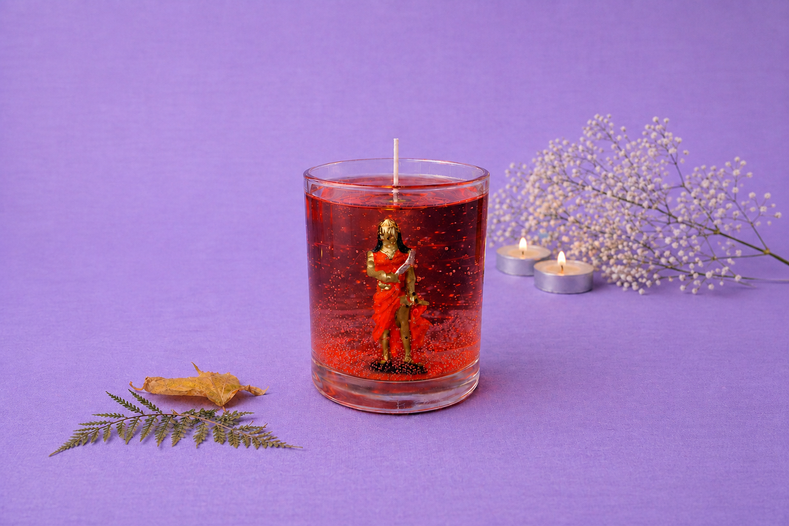 Vela em Gel Iansã artesanal em copo de vidro redondo com parafina em gel vermelha translúcida, contendo uma representação de Iansã em seu interior. A composição está sobre fundo lilás com flores delicadas, velas acesas e elementos naturais decorativos.