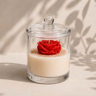 Vela Cúpula Rosa Vermelha artesanal em recipiente de vidro transparente com tampa de vidro refinada. A composição apresenta uma rosa vermelha esculpida em cera sobre uma base de cera clara, destacada por iluminação suave e sombras naturais de folhagens ao fundo. A imagem transmite elegância, delicadeza e um estilo decorativo sofisticado.
