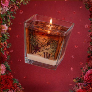 Vela aromática artesanal em recipiente quadrado de vidro com especiarias decorativas e chama acesa.