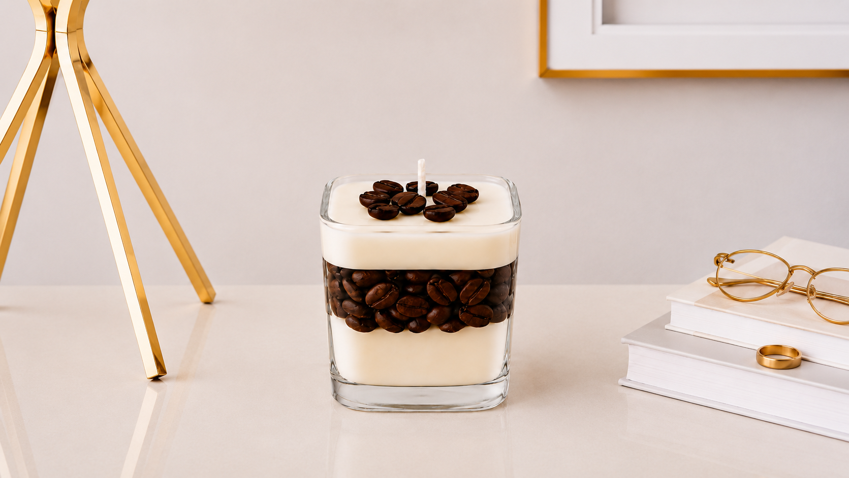 Vela Aromática Coffee artesanal em recipiente de vidro quadrado transparente, composta por camadas de cera creme e grãos de café decorativos ao centro e na superfície, apresentada em ambiente sofisticado com livros, óculos e detalhes dourados.