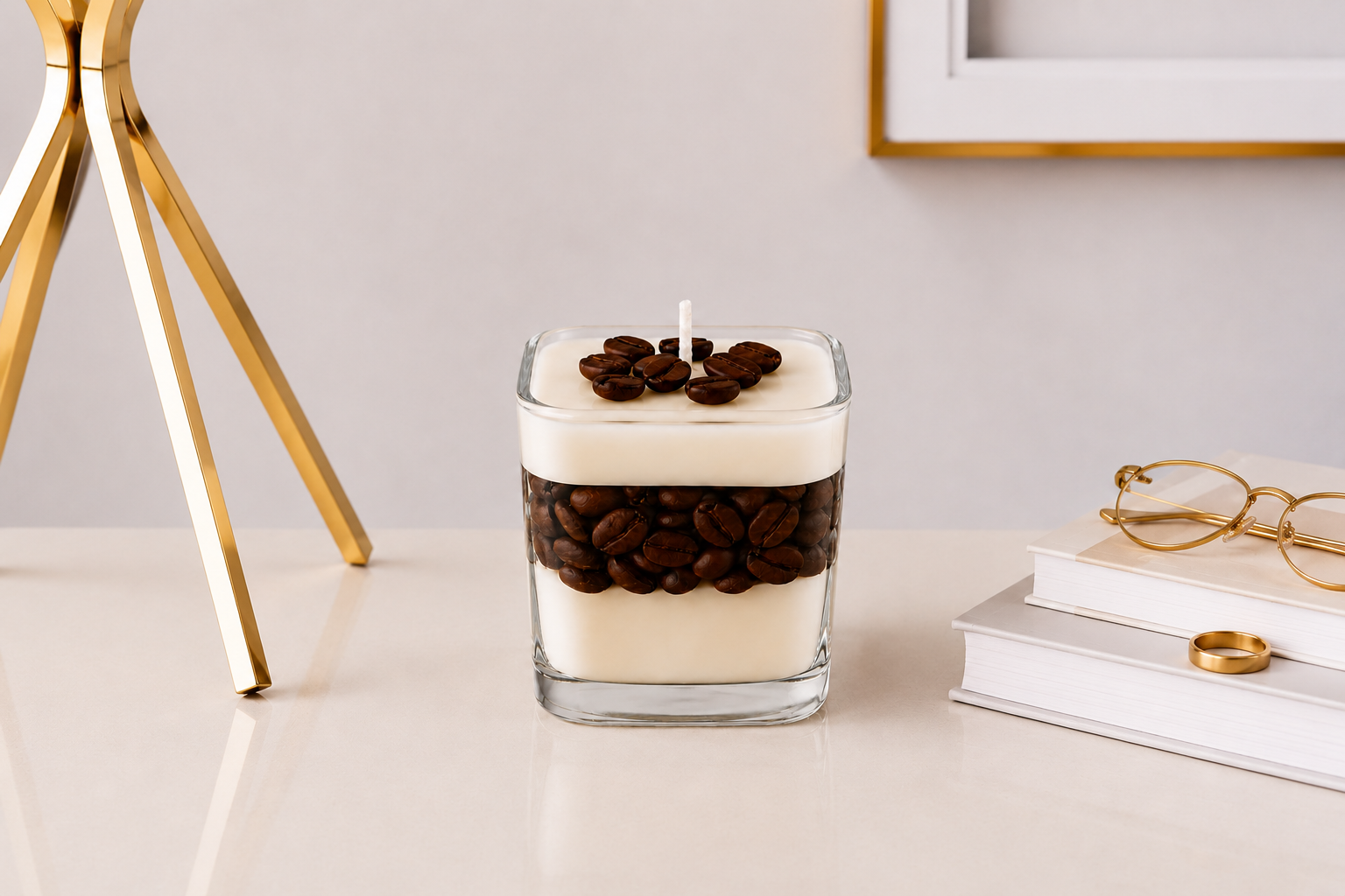 Vela Aromática Coffee artesanal em recipiente de vidro quadrado transparente, com camadas de cera creme e grãos de café decorativos, posicionada sobre uma mesa elegante ao lado de livros e objetos dourados em um ambiente sofisticado.