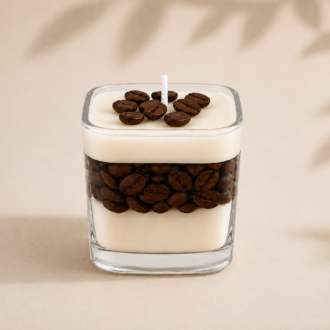 Vela Aromática Coffee artesanal em recipiente de vidro quadrado transparente, com camadas de cera creme e grãos de café decorativos, fotografada em fundo neutro com sombras suaves de folhas, destacando seu design elegante e minimalista.