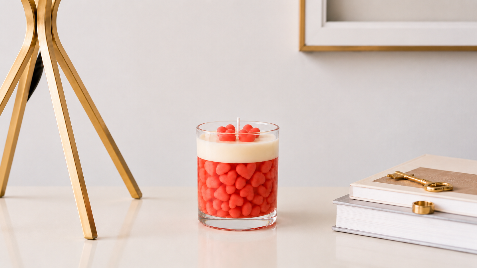 Vela aromática decorativa com pequenos corações em tons de rosa dentro de recipiente de vidro transparente, posicionada sobre uma mesa clara com livros e detalhes dourados em uma composição elegante e minimalista.