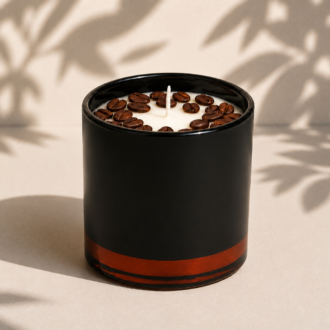 Vela Aromática de Café Torrado em recipiente preto fosco com base âmbar translúcida, decorada com grãos de café sobre a superfície da cera. A composição está posicionada em um fundo claro com sombras suaves de folhas, destacando o design elegante e a proposta sofisticada da fragrância inspirada no café recém-torrado.