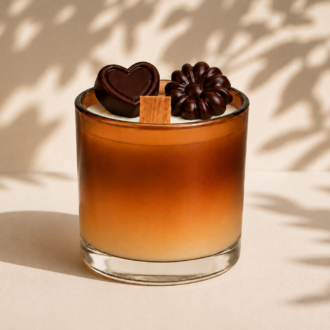 Vela Aromática de Chocolate em copo de vidro degradê nos tons de creme e marrom chocolate, decorada com miniaturas de chocolates em formato de coração e flor sobre a superfície. A composição apresenta sombras suaves de folhas ao fundo, criando uma atmosfera elegante, aconchegante e sofisticada.