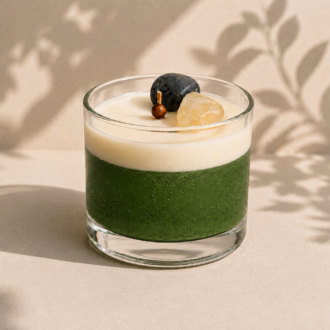 Vela Aromática de Cristais em copo de vidro transparente, com camada inferior verde translúcida e camada superior cremosa. A decoração inclui cristais naturais e elementos ornamentais sobre a superfície da vela. A composição apresenta iluminação suave, sombras orgânicas de folhagens ao fundo e uma estética minimalista e sofisticada.