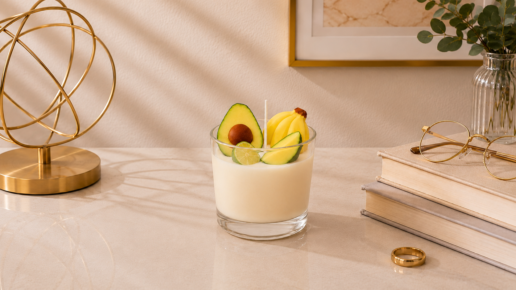 Vela Aromática de Frutas Tropicais em copo de vidro transparente, decorada com miniaturas de frutas tropicais, incluindo abacate, banana e limão. A vela está posicionada sobre uma superfície clara em um ambiente sofisticado, com livros, objeto decorativo dourado, vaso com folhagens e iluminação natural suave, criando uma atmosfera elegante e acolhedora.
