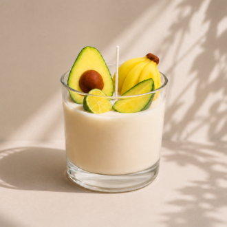 Vela Aromática de Frutas Tropicais em copo de vidro transparente, decorada com miniaturas de abacate, banana e limão sobre a superfície da cera. A composição está posicionada em um cenário minimalista com fundo bege suave, iluminação natural e sombras delicadas de folhagens, transmitindo frescor, leveza e inspiração tropical.