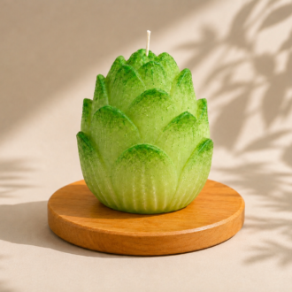 Vela Aromática Flor de Lótus artesanal em formato de flor, com pétalas em tons suaves de verde e acabamento texturizado. A peça está posicionada sobre uma base redonda de madeira, com iluminação natural suave e sombras orgânicas de folhas ao fundo, transmitindo uma atmosfera de serenidade, equilíbrio e conexão com a natureza.