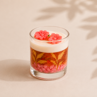Vela Aromática Floral Rosé artesanal em copo de vidro transparente, com camada superior de parafina branca, gel decorativo em tom rosé com folhas douradas ornamentais, pedras decorativas rosadas e flores delicadas na superfície, apresentada sobre fundo claro com sombras suaves de folhagens.