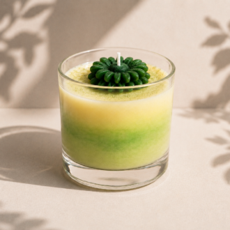 Vela Aromática Flower Green em copo de vidro transparente, com efeito marmorizado em suaves tons de verde e creme. A vela apresenta uma delicada flor verde esculpida em cera no topo, posicionada em um cenário minimalista de fundo claro com sombras naturais de folhagens, transmitindo frescor, serenidade e elegância.