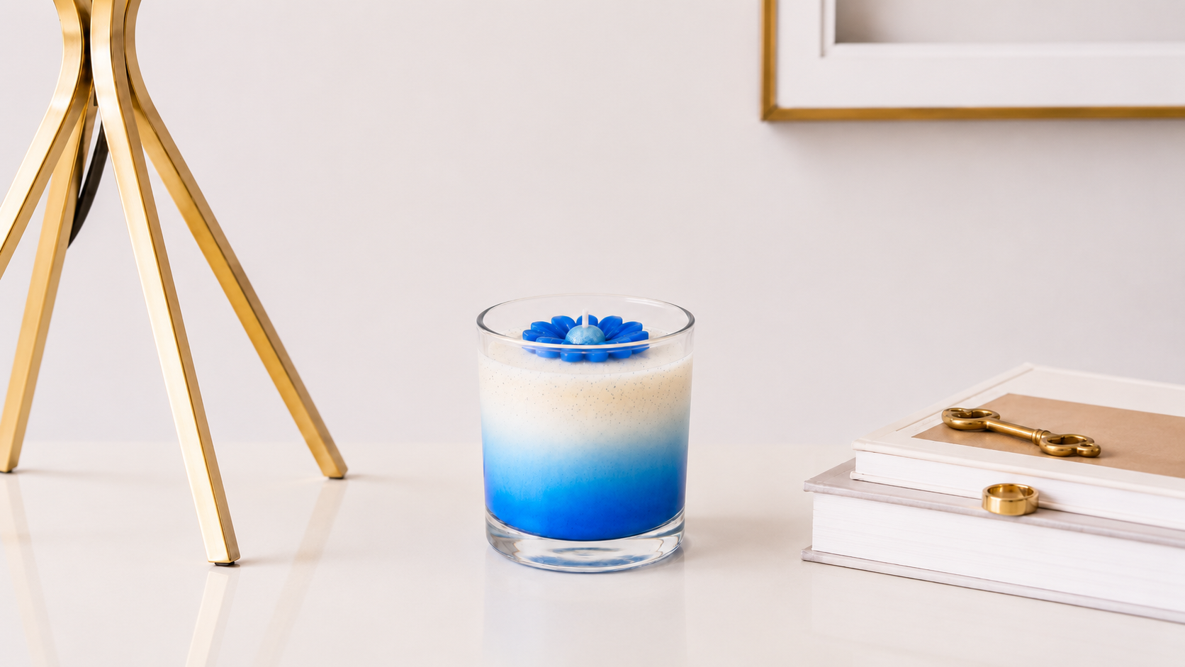 Vela aromática em vidro transparente com degradê em tons de azul intenso e branco, decorada com uma margarida azul esculpida em cera, apresentada em ambiente sofisticado com elementos dourados e livros decorativos ao fundo.