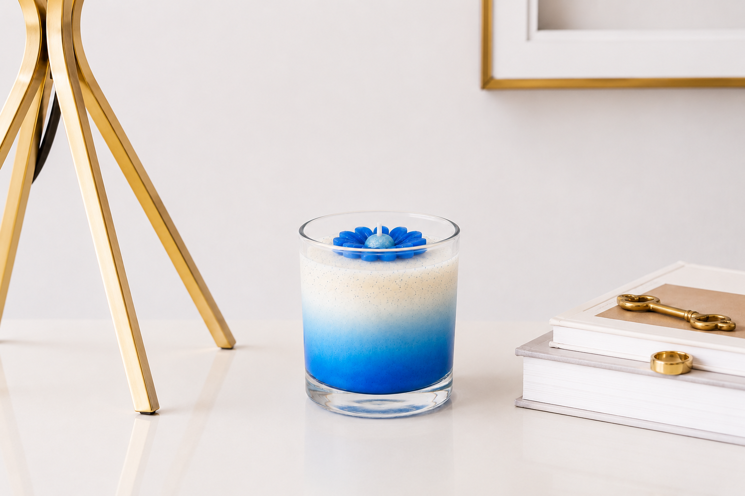 Vela aromática em vidro transparente com degradê em tons de azul e branco, decorada com uma margarida azul esculpida em cera, apresentada em ambiente sofisticado com luminária dourada, livros decorativos e acessórios em acabamento dourado.