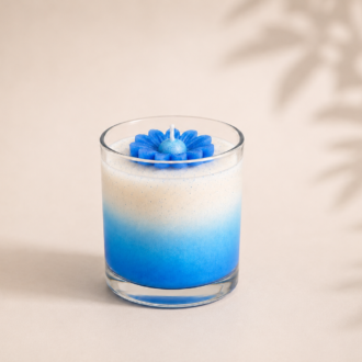 Vela aromática em vidro transparente com degradê em tons de azul e branco, decorada com uma margarida azul esculpida em cera, posicionada sobre fundo bege suave com sombras delicadas de folhas, em composição minimalista e elegante.