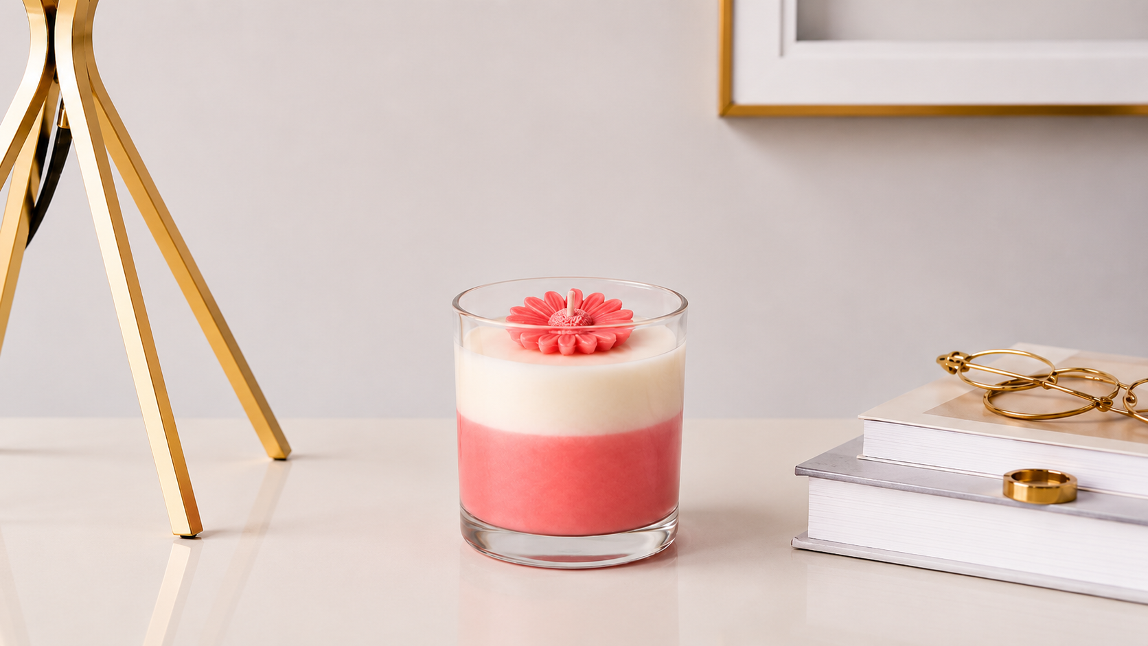 Vela Aromática Margarida Rosé em copo de vidro transparente, com camadas em tons rosé e branco e delicada margarida decorativa na superfície, apresentada em ambiente sofisticado com livros e elementos dourados.