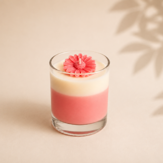 Vela Aromática Margarida Rosé em copo de vidro transparente, com camadas em tons rosé e branco e margarida esculpida em cera no topo, apresentada sobre fundo bege suave com delicadas sombras de folhas ao fundo.