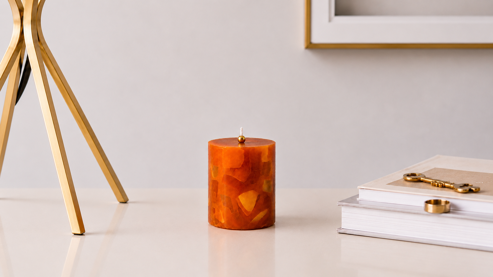 Vela aromática cilíndrica em tom âmbar com efeito mosaico translúcido, apresentada em ambiente sofisticado com luminária dourada, livros decorativos e detalhes em dourado, destacando seu visual elegante e artesanal.