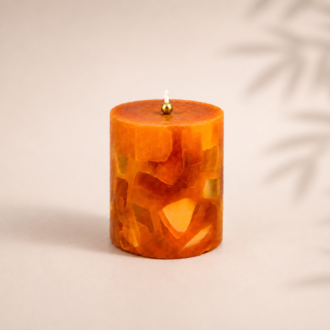 Vela aromática mosaico em tons âmbar e dourado, com acabamento artesanal em peças translúcidas, fotografada sobre fundo neutro com delicadas sombras botânicas ao fundo.
