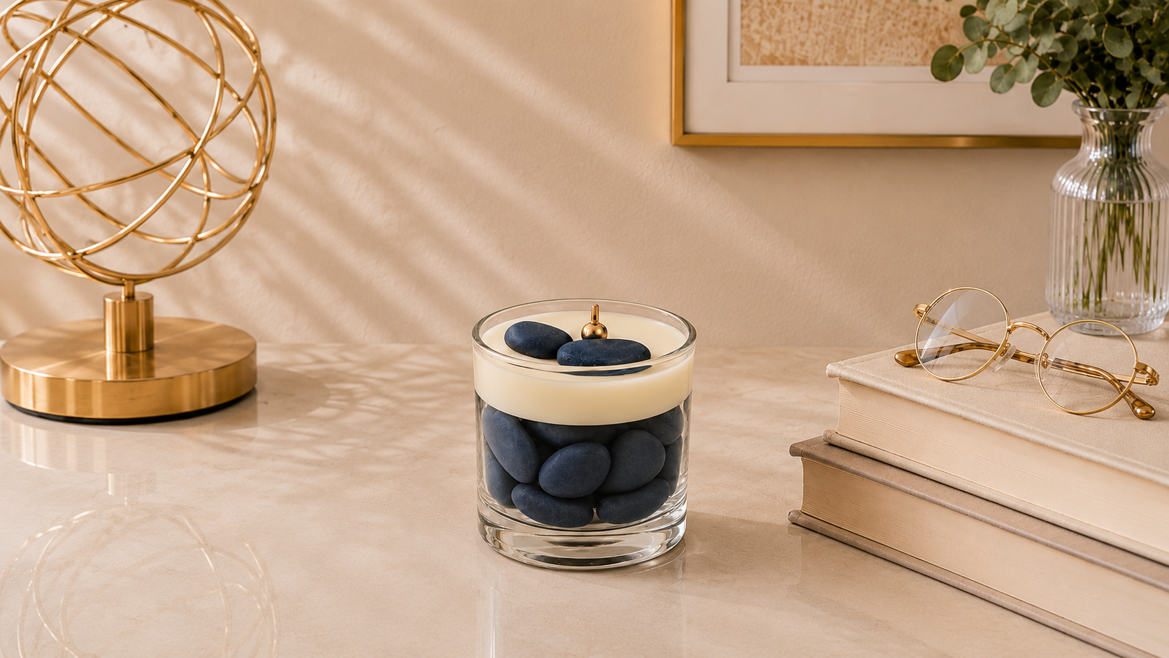 Vela Aromática Quartzo Azul em recipiente de vidro transparente, preenchido com pedras decorativas em tom azul profundo e finalizada com uma camada superior de cera cremosa. A vela possui pavio metálico dourado centralizado e está posicionada sobre uma elegante superfície clara. Ao redor da composição aparecem livros em tons neutros, óculos de armação dourada, objeto decorativo metálico e folhagens delicadas, criando uma atmosfera sofisticada, acolhedora e contemporânea.