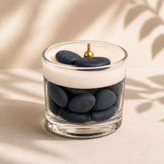 Vela Aromática Quartzo Azul em recipiente de vidro transparente, composta por pedras decorativas azul-escuras e camada superior de cera branca cremosa. A vela apresenta pavio metálico dourado centralizado e duas pedras azuis decorativas sobre a superfície. A composição está fotografada em fundo bege claro com sombras suaves de folhagens projetadas ao fundo, criando uma atmosfera elegante, serena e sofisticada.