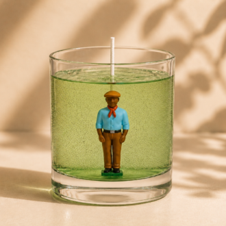 Vela em Gel Boiadeiro artesanal em recipiente de vidro transparente com gel verde, contendo a figura de um Boiadeiro em seu interior, fotografada sobre fundo bege suave com sombras naturais de folhas em composição minimalista e elegante.