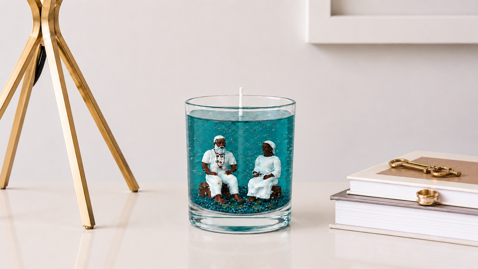 Vela em gel transparente azul com miniaturas artesanais de um casal de Pretos Velhos em seu interior, posicionada sobre uma mesa clara ao lado de livros e elementos dourados em um ambiente sofisticado e acolhedor.