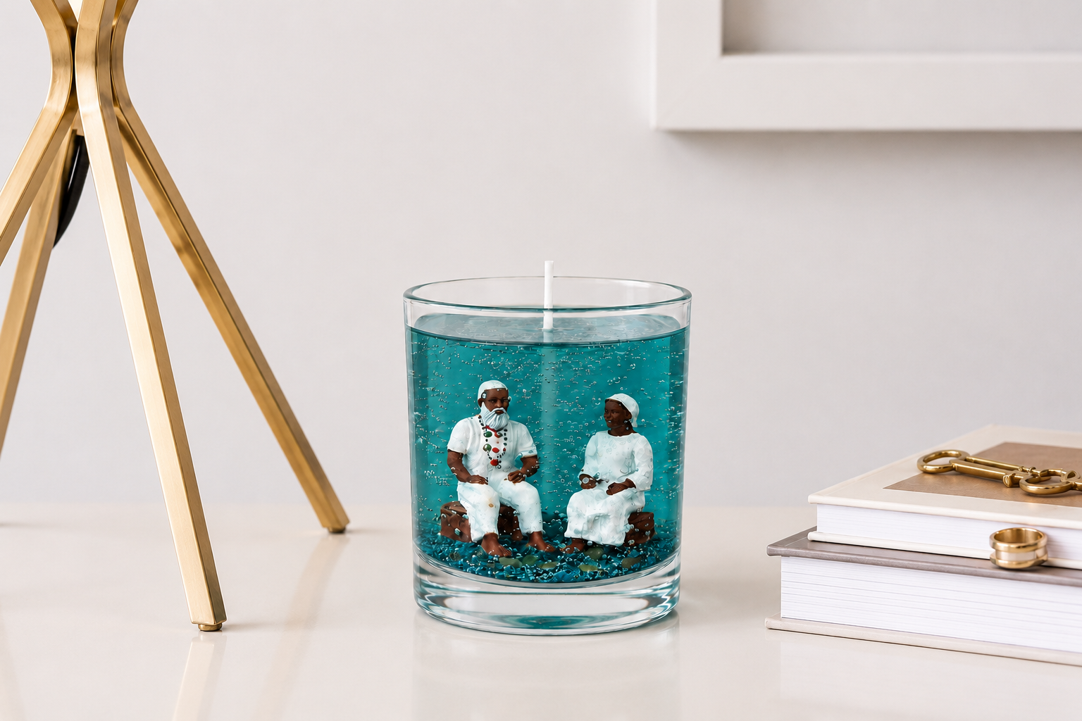 Vela em gel azul-turquesa com miniaturas artesanais de um casal de Pretos Velhos em seu interior, apresentada em um ambiente sofisticado com decoração minimalista, livros e detalhes dourados ao fundo.