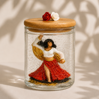 Vela artesanal em gel transparente com figura de Cigana dançando, vestida com saia vermelha e blusa branca com detalhes dourados, acomodada em recipiente de vidro com tampa de madeira decorada por duas rosas. A composição apresenta iluminação suave, sombras orgânicas e detalhes dourados no interior da peça, criando um visual elegante e acolhedor.