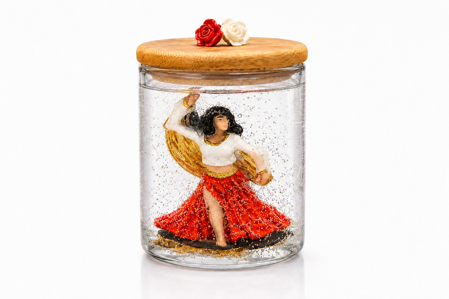 Vela artesanal em gel transparente com escultura de Cigana dançando, vestida com saia vermelha e blusa branca com detalhes dourados. A peça está acondicionada em um elegante recipiente de vidro com tampa de madeira decorada por duas rosas, uma vermelha e uma branca, destacando a delicadeza dos detalhes e a transparência do gel.