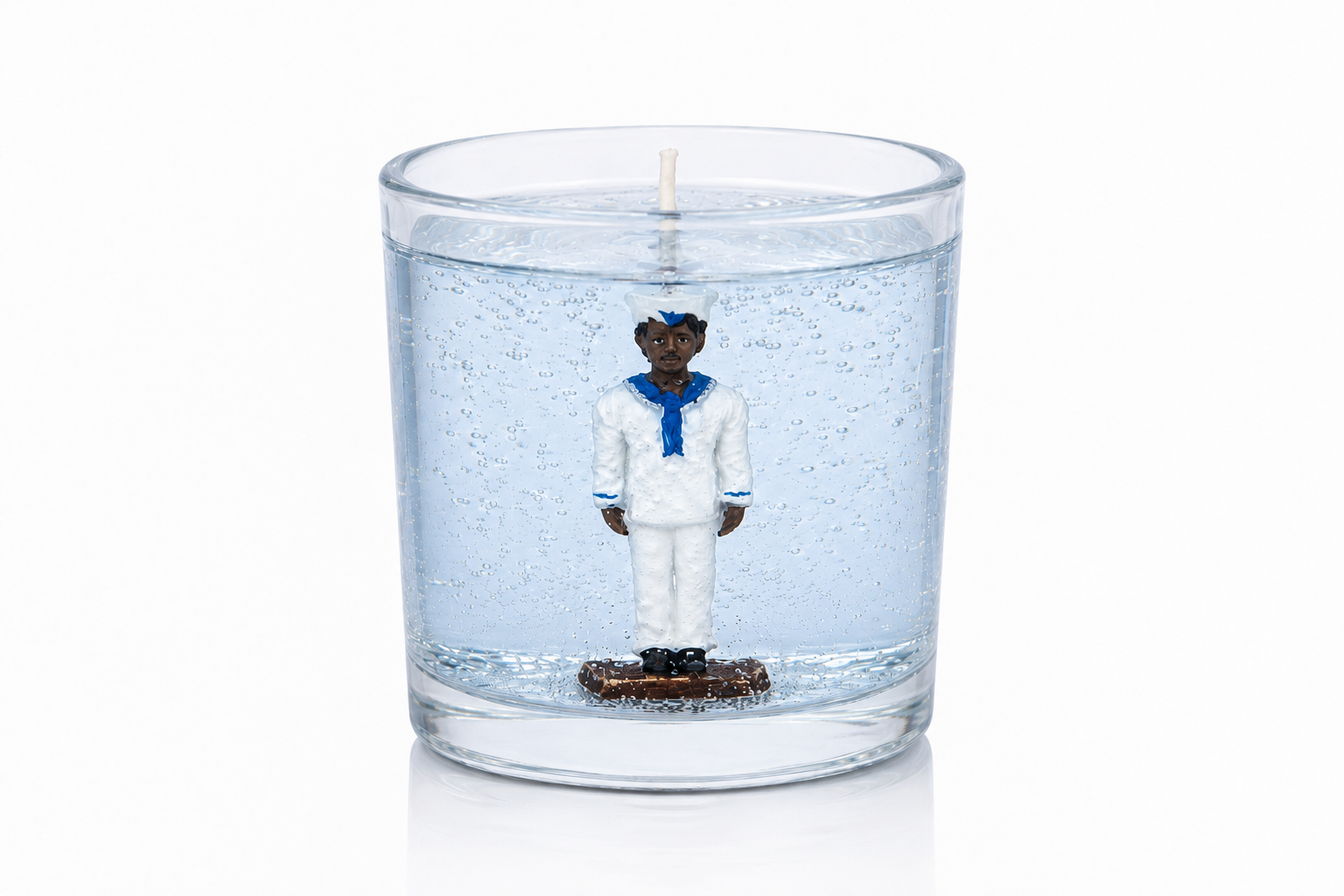 Vela artesanal em gel azul-claro transparente com figura de Marinheiro em uniforme branco e detalhes azuis posicionada no centro do recipiente de vidro cilíndrico. A composição destaca a transparência do gel, as pequenas bolhas decorativas e a estética marítima associada à proteção, serenidade e equilíbrio.