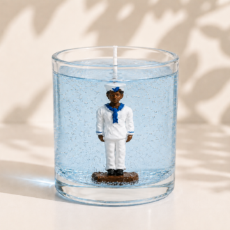 Vela artesanal em gel transparente azul-claro com figura de Marinheiro vestindo uniforme branco com detalhes azuis, posicionada no centro de um recipiente de vidro cilíndrico. A composição apresenta iluminação suave e sombras orgânicas ao fundo, transmitindo serenidade, leveza e inspiração nas águas do mar.