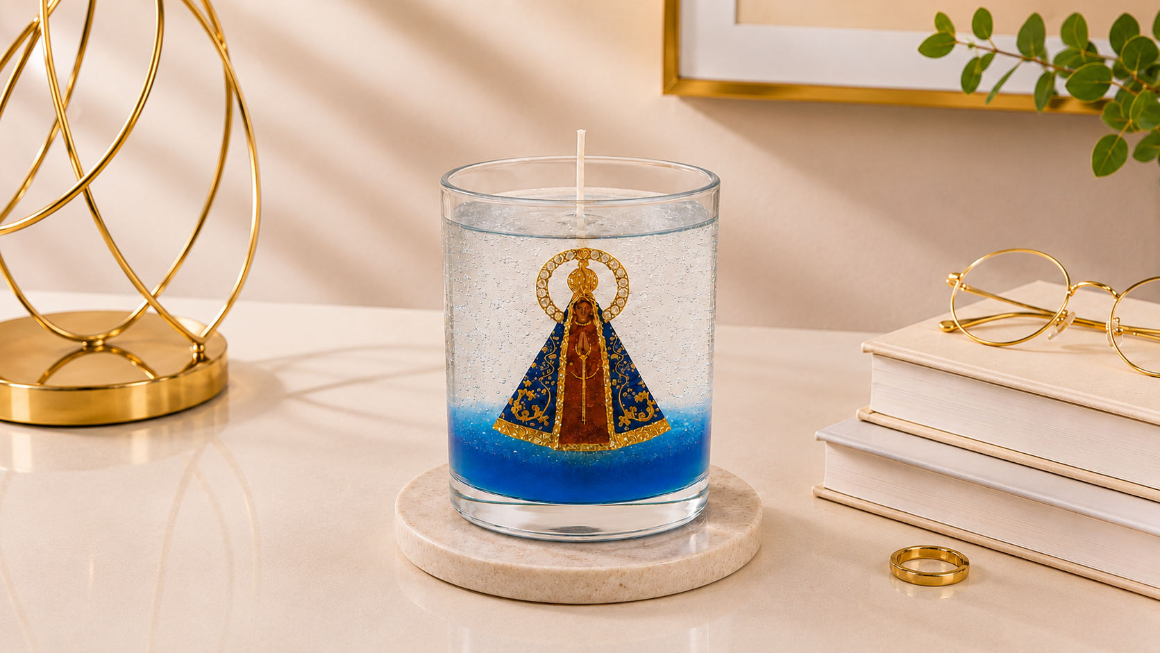 Vela em Gel Nossa Senhora Aparecida artesanal em copo de vidro transparente, com imagem central da Padroeira do Brasil envolvida por parafina em gel cristalina e delicados detalhes em azul na base. A vela está posicionada sobre uma base clara em um ambiente elegante, com objeto decorativo dourado, livros neutros, óculos e iluminação suave, transmitindo serenidade, fé e sofisticação.
