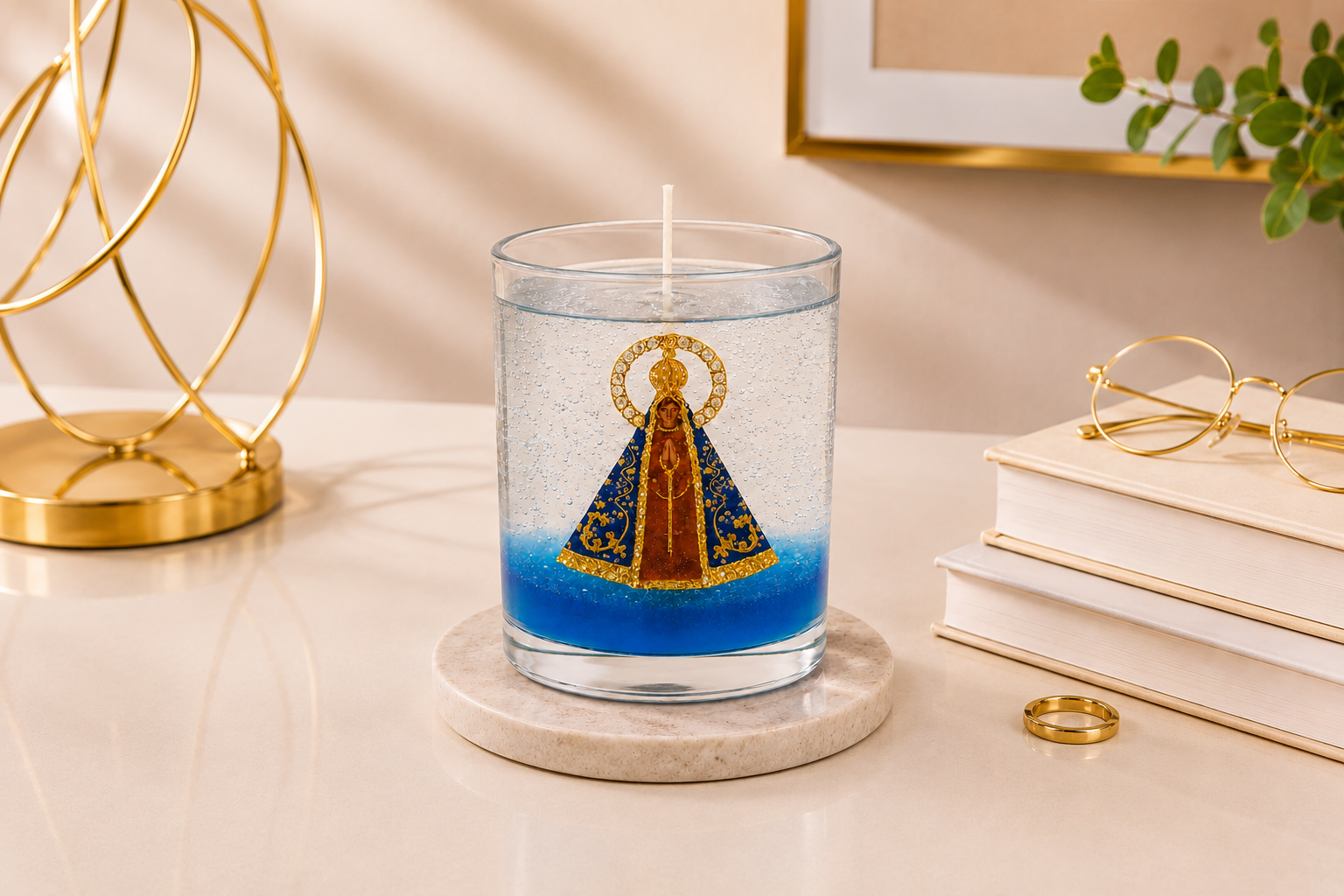 Vela em Gel Nossa Senhora Aparecida artesanal em copo de vidro transparente, com imagem central da Padroeira do Brasil envolvida por parafina em gel cristalina e delicados detalhes em azul na base. A vela está posicionada sobre uma base clara em um ambiente elegante, com objeto decorativo dourado, livros neutros, óculos e iluminação suave, transmitindo serenidade, fé e sofisticação.