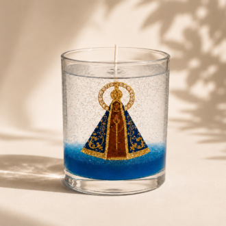 Vela em Gel Nossa Senhora Aparecida artesanal em copo de vidro transparente, com imagem central da Padroeira do Brasil envolvida por parafina em gel cristalina e delicadas bolhas decorativas. A composição apresenta uma elegante base azul que destaca a imagem religiosa e transmite serenidade, fé e devoção. A vela está posicionada em um cenário minimalista de tons claros, com iluminação suave e sombras naturais de folhagens ao fundo, criando uma atmosfera sofisticada e acolhedora.