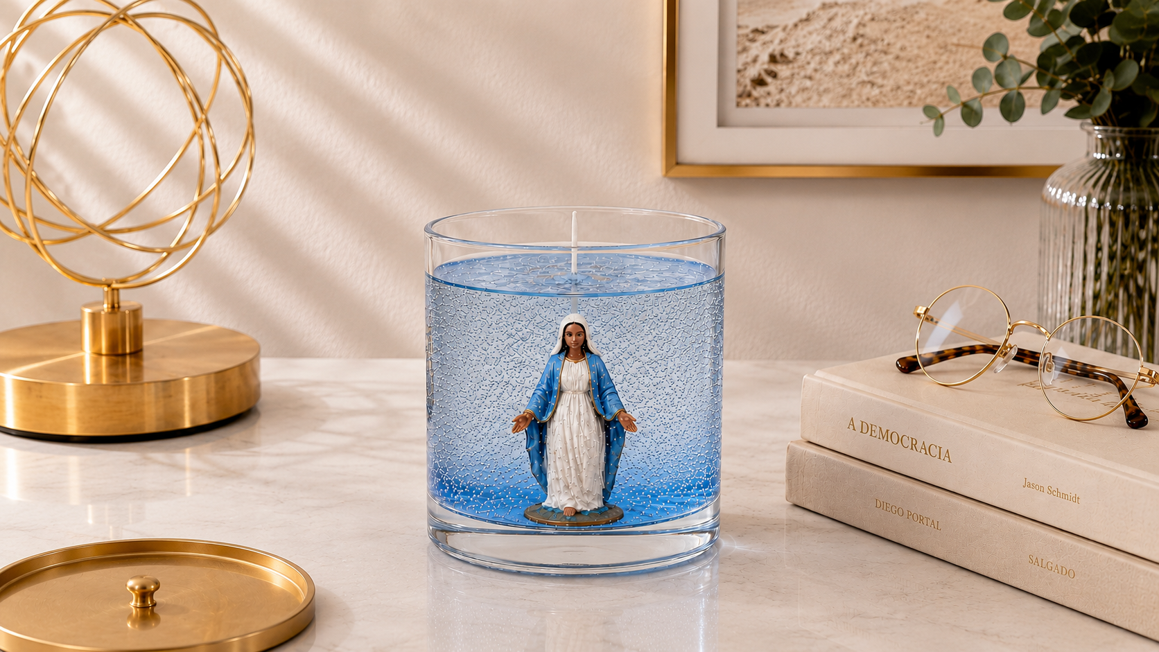 Vela em Gel Nossa Senhora das Graças artesanal em recipiente de vidro transparente com gel azul, apresentando a imagem de Nossa Senhora das Graças em seu interior, fotografada em ambiente sofisticado com livros decorativos, elementos dourados e iluminação natural suave.