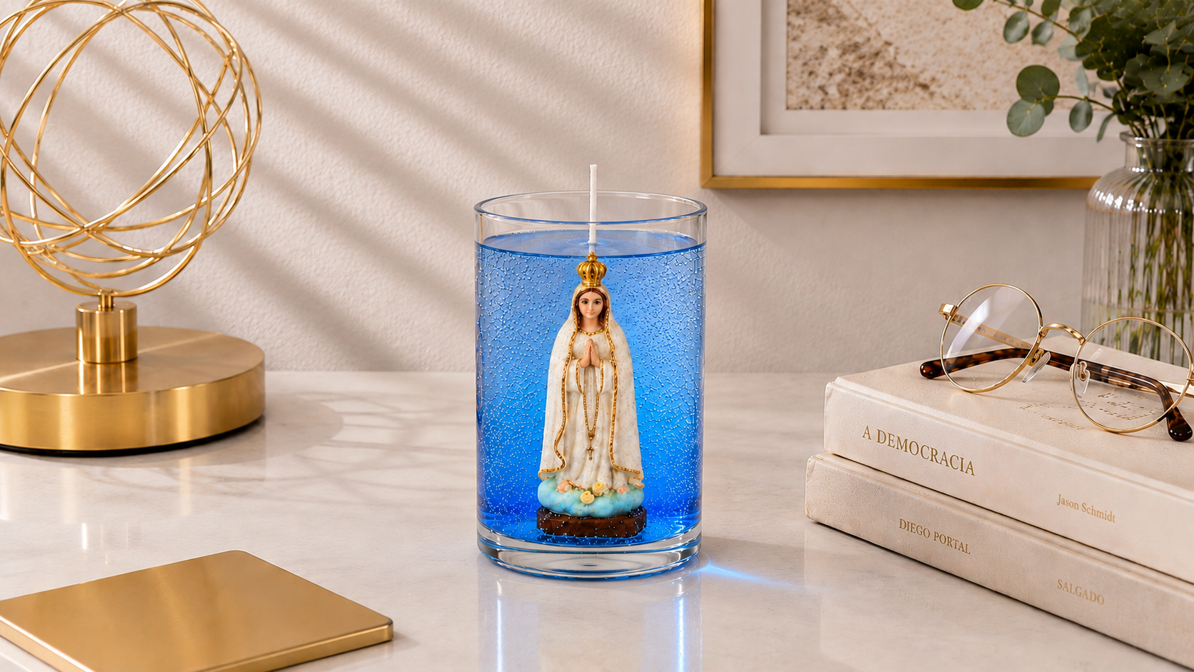 Vela em gel azul transparente com microbolhas decorativas em recipiente cilíndrico de vidro. No interior encontra-se uma imagem de Nossa Senhora de Fátima com vestes brancas, detalhes dourados e mãos unidas em oração. A peça está posicionada sobre uma superfície clara em ambiente sofisticado com livros, óculos, vaso decorativo e objeto dourado ao fundo, transmitindo elegância, espiritualidade e devoção.