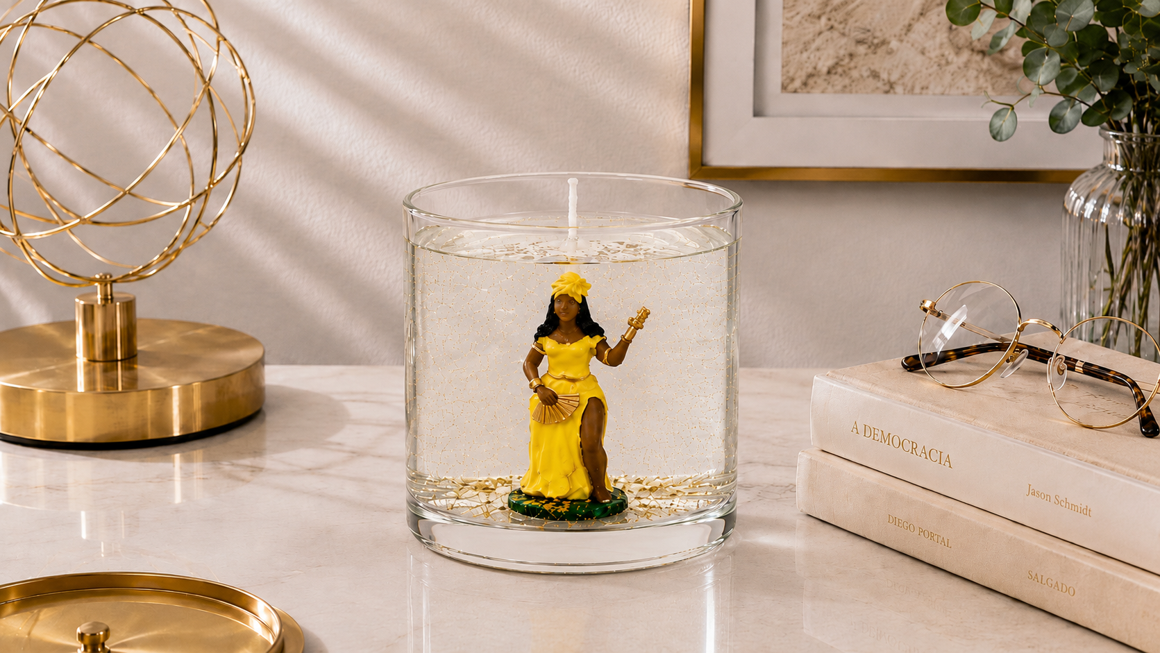 Vela em gel transparente com imagem de Oxum vestida de amarelo dentro de recipiente cilíndrico de vidro. A peça está posicionada sobre uma superfície clara em ambiente sofisticado com luminária dourada, livros, óculos e elementos decorativos ao fundo, transmitindo uma atmosfera de prosperidade, beleza e serenidade.