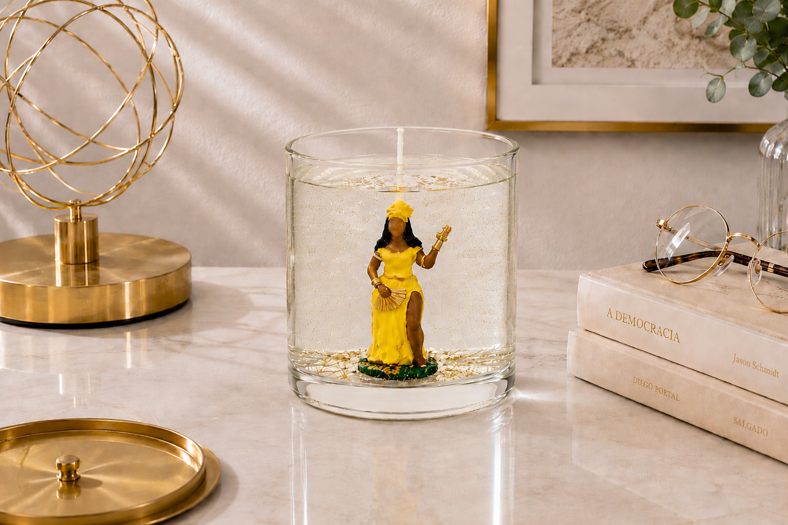 Vela artesanal em gel transparente com imagem de Oxum vestida de amarelo no interior de um recipiente de vidro cilíndrico. A peça está posicionada sobre uma bancada elegante ao lado de livros decorativos, óculos e objetos dourados, criando uma atmosfera sofisticada que remete à prosperidade, beleza e harmonia.